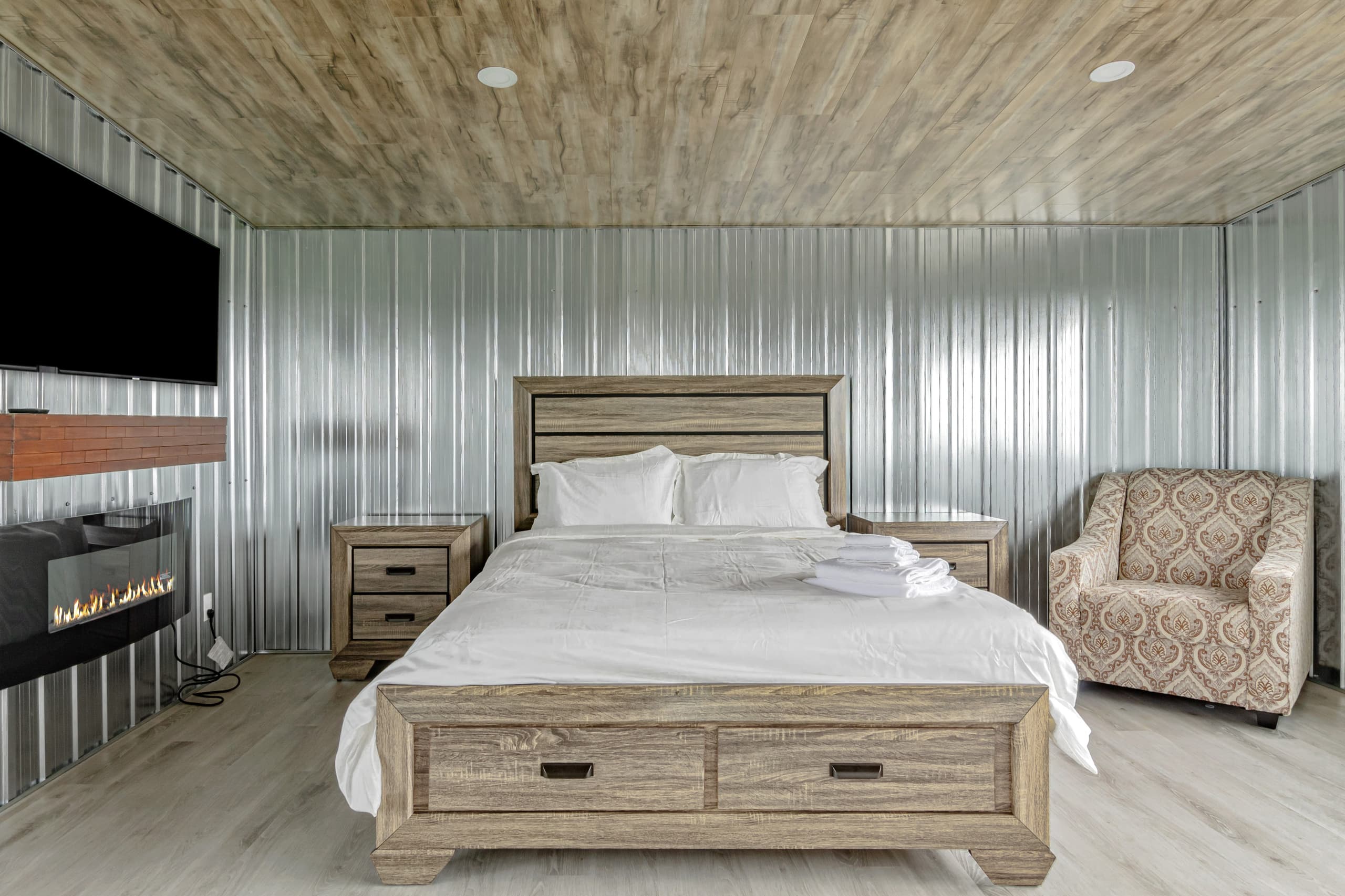 A bedroom lined in corrugated galvanised steel with a fireplace, television and timber mantel opposite the bed