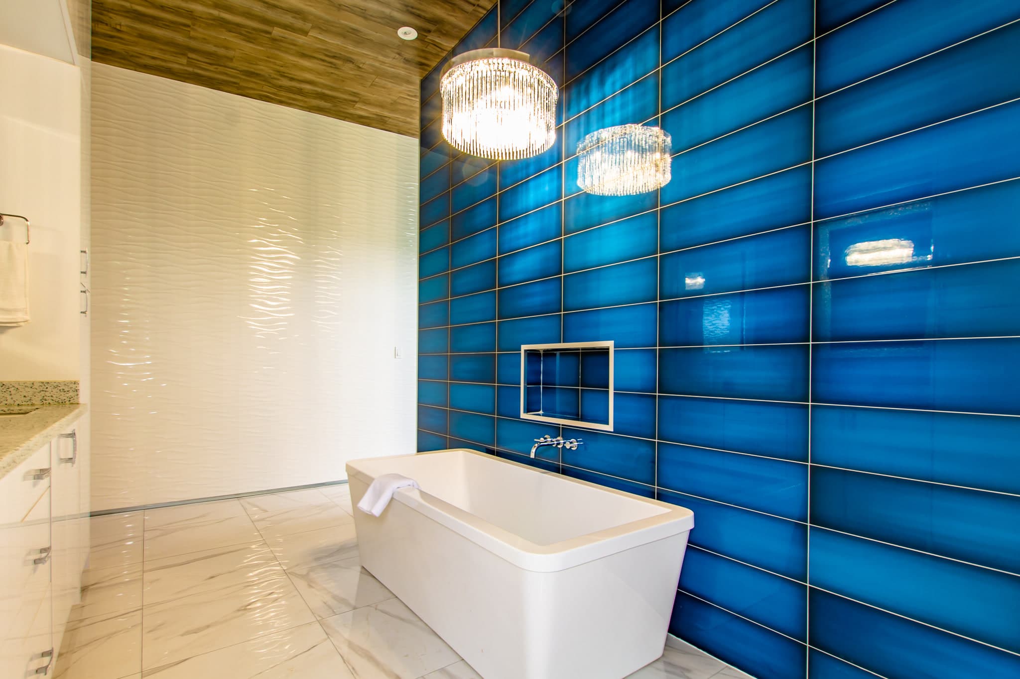 A freestanding soaker tub in front of a floor-to-ceiling wall of deep blue glazed tile, lit by two crystal pendants